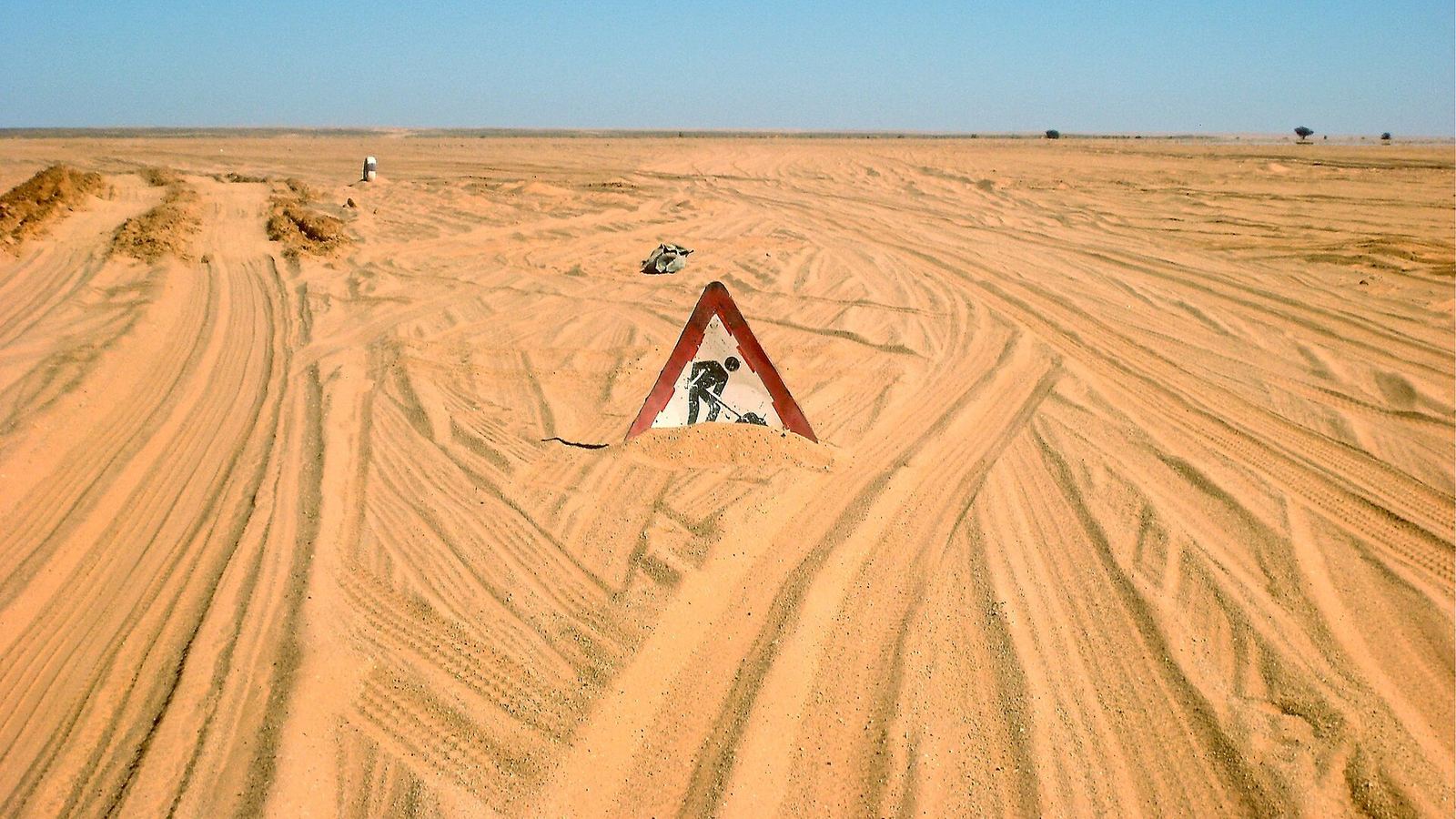 Sand tracks across the Libyan desert with a half-buried roadworks sign