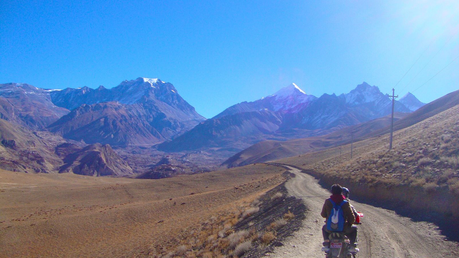 Motorcycle with two riders (seen from behind) on a dirt road in the arid Mustang valley with snow-capped Himalayan peaks under a deep blue sky