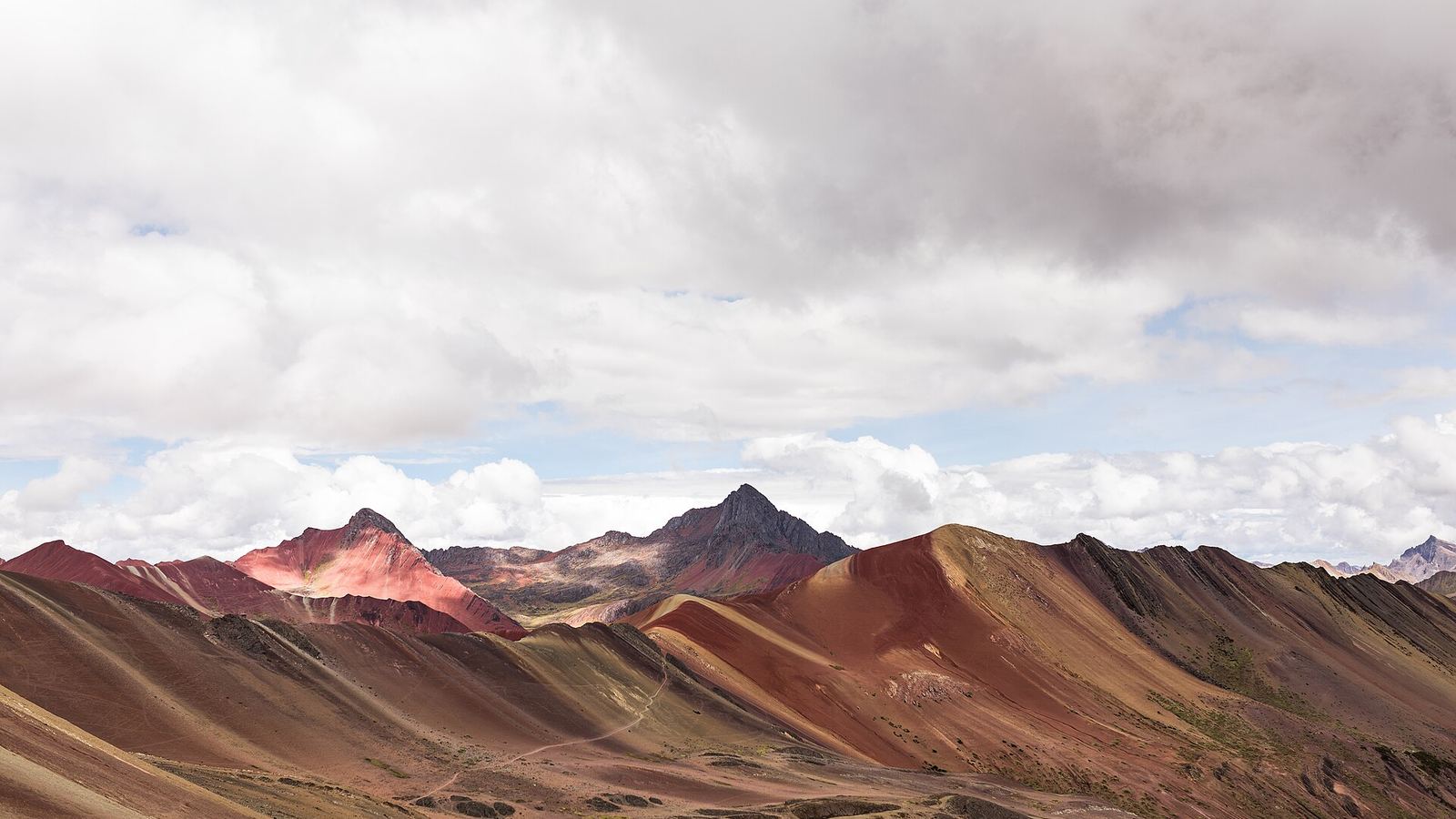 Vinicunca (Rainbow Mountain) in the Cusco region, striped red, ochre and green ridges under heavy cloud, a thin trail across the slope.