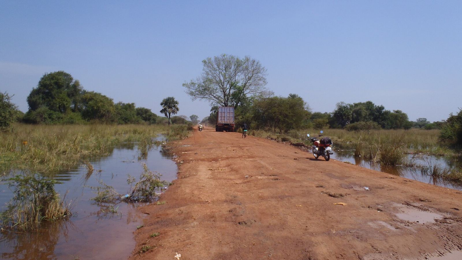 Flooded red dirt road outside Yirol Town, South Sudan, with a distant truck and motorbikes