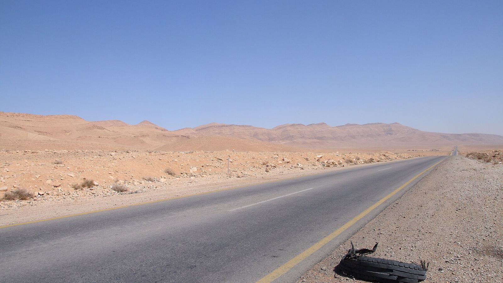 Empty paved desert road toward barren hills with a worn-out tyre on the shoulder, taken in March 2009