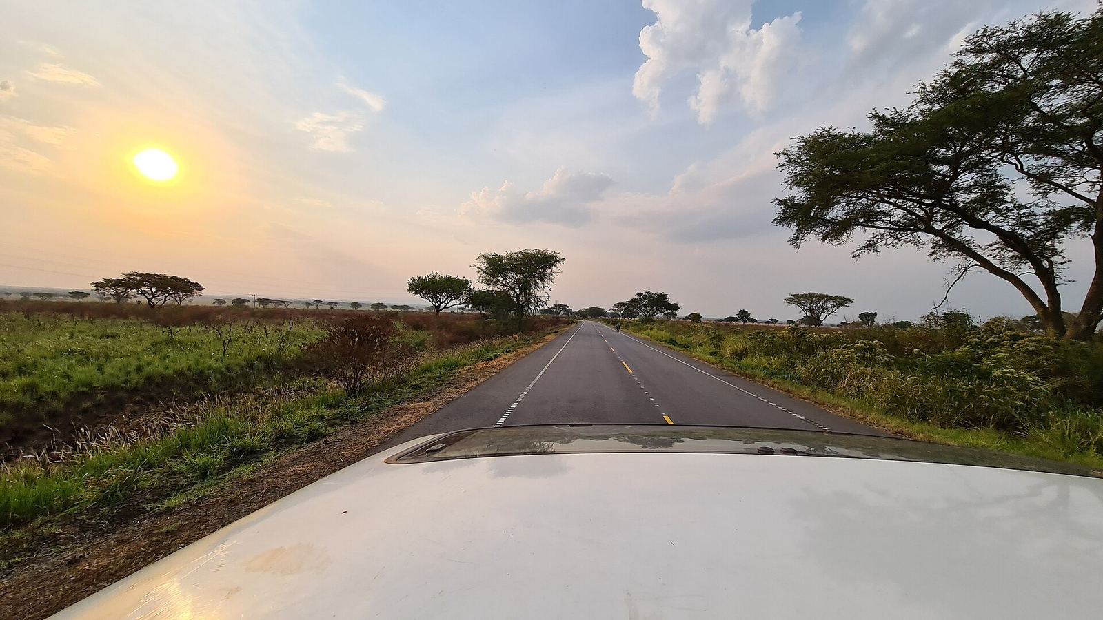 Sunset over an empty highway lined with acacias through Queen Elizabeth National Park, seen over the bonnet of a white vehicle
